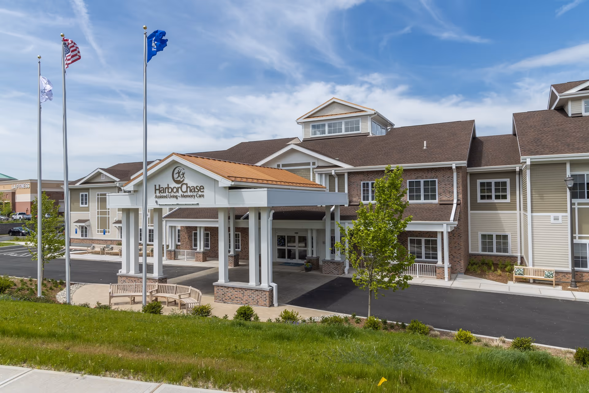 Exterior view of HarborChase Assisted Living and Memory Care building with a covered entrance, three flagpoles with flags, green lawn, and a partly cloudy sky.