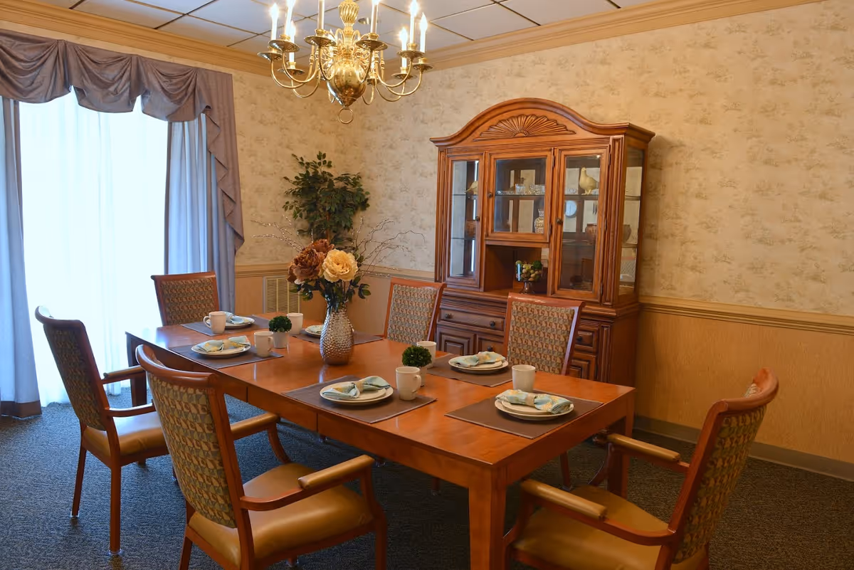A traditional dining room with a wooden table set for six, a china cabinet, chandelier, and floral centerpiece.