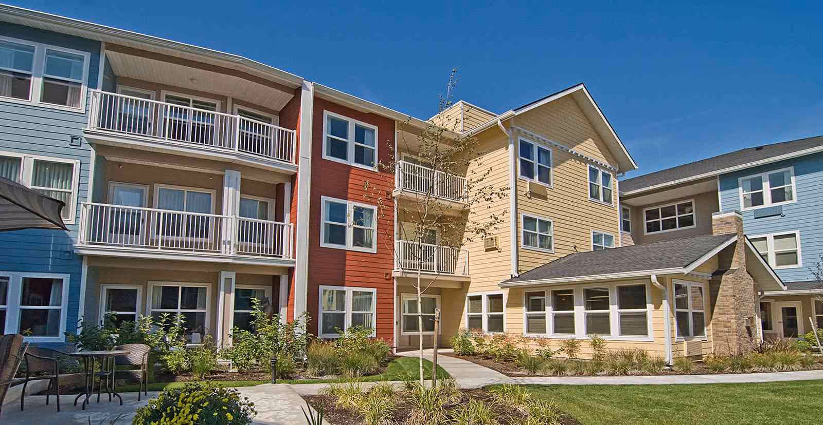 Exterior view of a colorful three-story senior living building with balconies, landscaped courtyard, and outdoor seating.