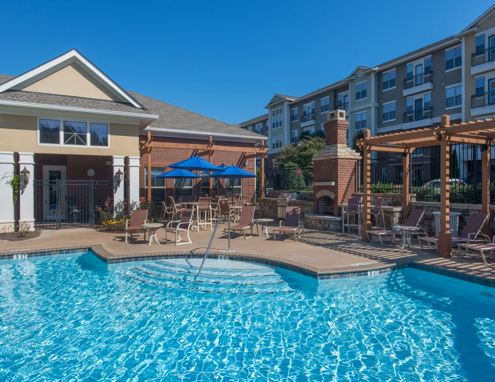 Outdoor swimming pool area with clear blue water, surrounded by lounge chairs, tables with blue umbrellas, a brick fireplace, and pergolas. A multi-story residential building is visible in the background under a clear blue sky.