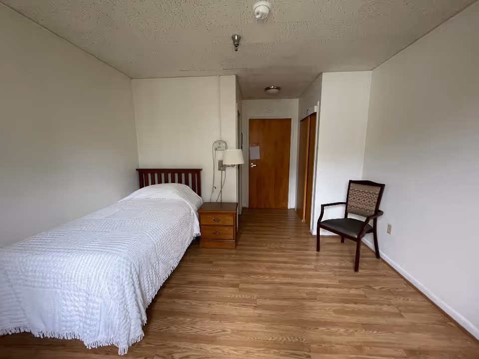 A simple bedroom with a single bed covered in a white textured bedspread, a wooden nightstand with a lamp, a wooden chair with armrests, and a wooden door at the far end. The room has light-colored walls and wood flooring.