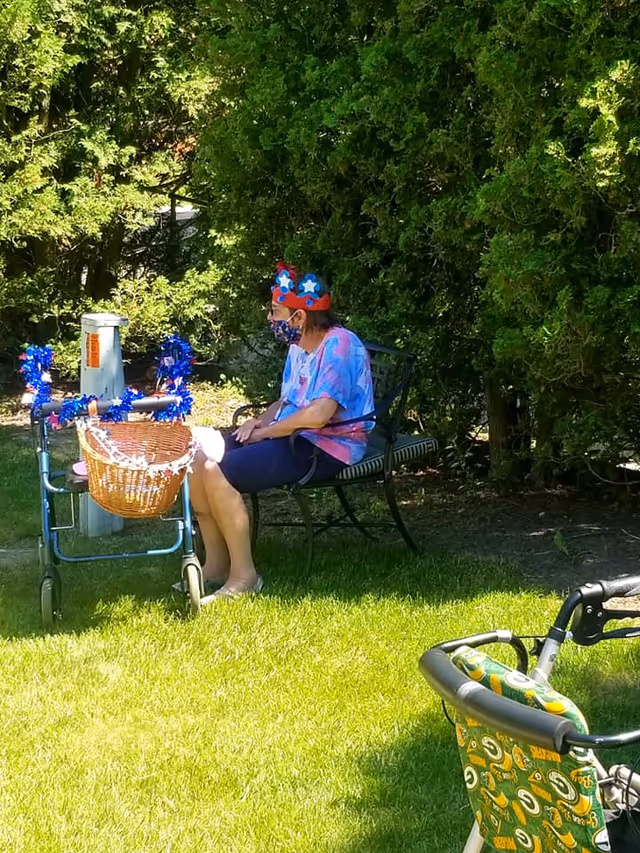 An elderly woman wearing a colorful crown and a face mask sits on a bench in a garden area next to a decorated walker with a basket. The scene is outdoors with green grass and dense bushes in the background. Another walker with a Green Bay Packers-themed seat is partially visible in the foreground.