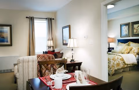 Interior view of a senior living facility showing a small dining table set for two with white dishes and napkins, a cozy living area with a striped armchair and a side table with a lamp, and a bedroom visible through an open doorway featuring a bed with yellow and white bedding and framed artwork above the headboard.
