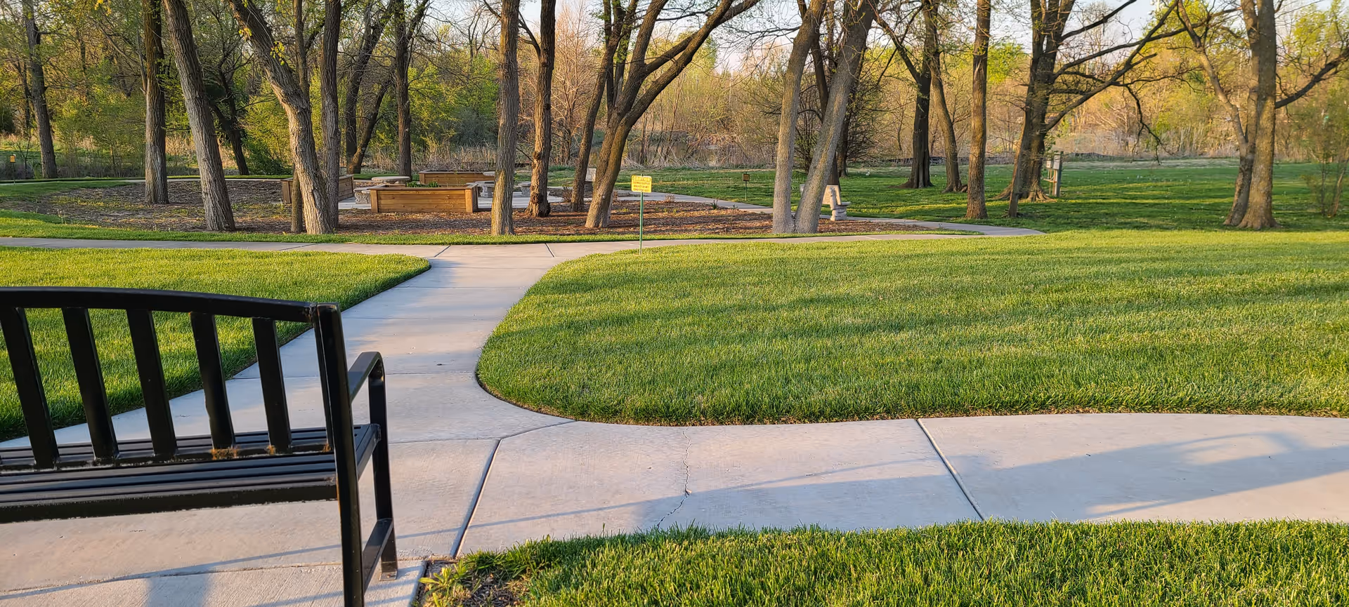 A peaceful outdoor park area with a black metal bench in the foreground, concrete walkways, well-maintained green grass, and several trees. In the background, there is a circular seating area with wooden benches surrounded by trees.