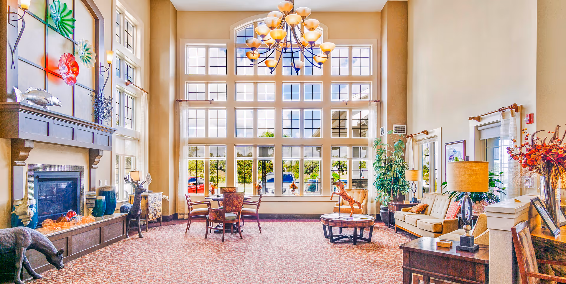 Sunlit community lounge with tall multi-pane windows, chandelier, seating areas, a fireplace, and decorative furnishings.