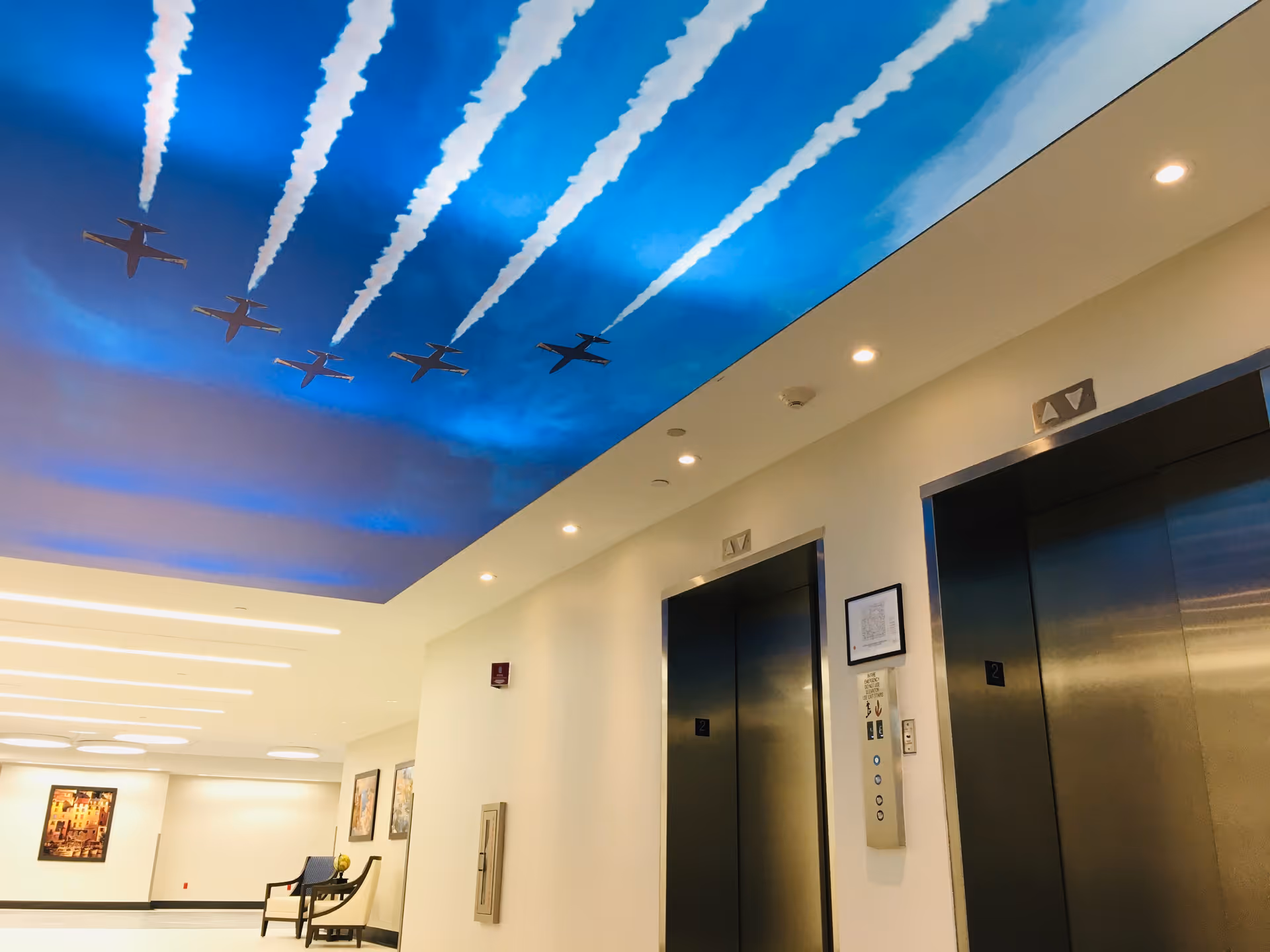 Interior hallway with two closed stainless steel elevators on the right side and a ceiling mural depicting six airplanes flying in formation with white smoke trails against a blue sky.