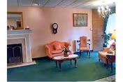A cozy living room with green carpet, a fireplace on the left, a small pink loveseat, two wooden chairs, a coffee table with a flower arrangement, a chandelier, and a framed painting on the wall.