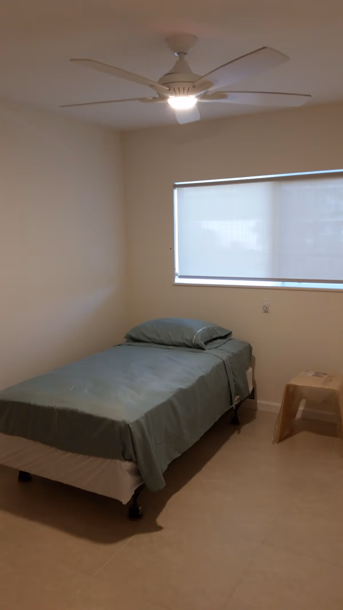 A simple bedroom with a single bed covered in green bedding, a matching pillow, a ceiling fan with a light, a window with a closed blind, and a small plastic stool beside the bed.