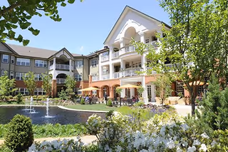 Exterior view of The Gatesworth senior living facility showing a multi-story building with balconies overlooking a landscaped garden area with a pond and water fountains, surrounded by trees and flowering plants under a clear blue sky.