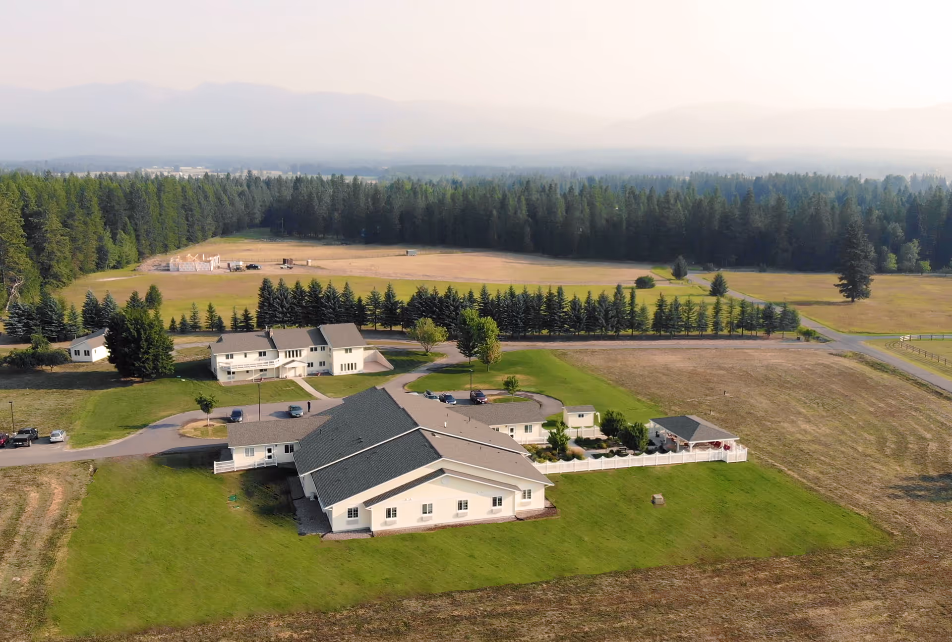 Aerial view of a rural memory-care campus with several white buildings, green lawns, a fenced yard, surrounding fields, tree line and distant mountains.