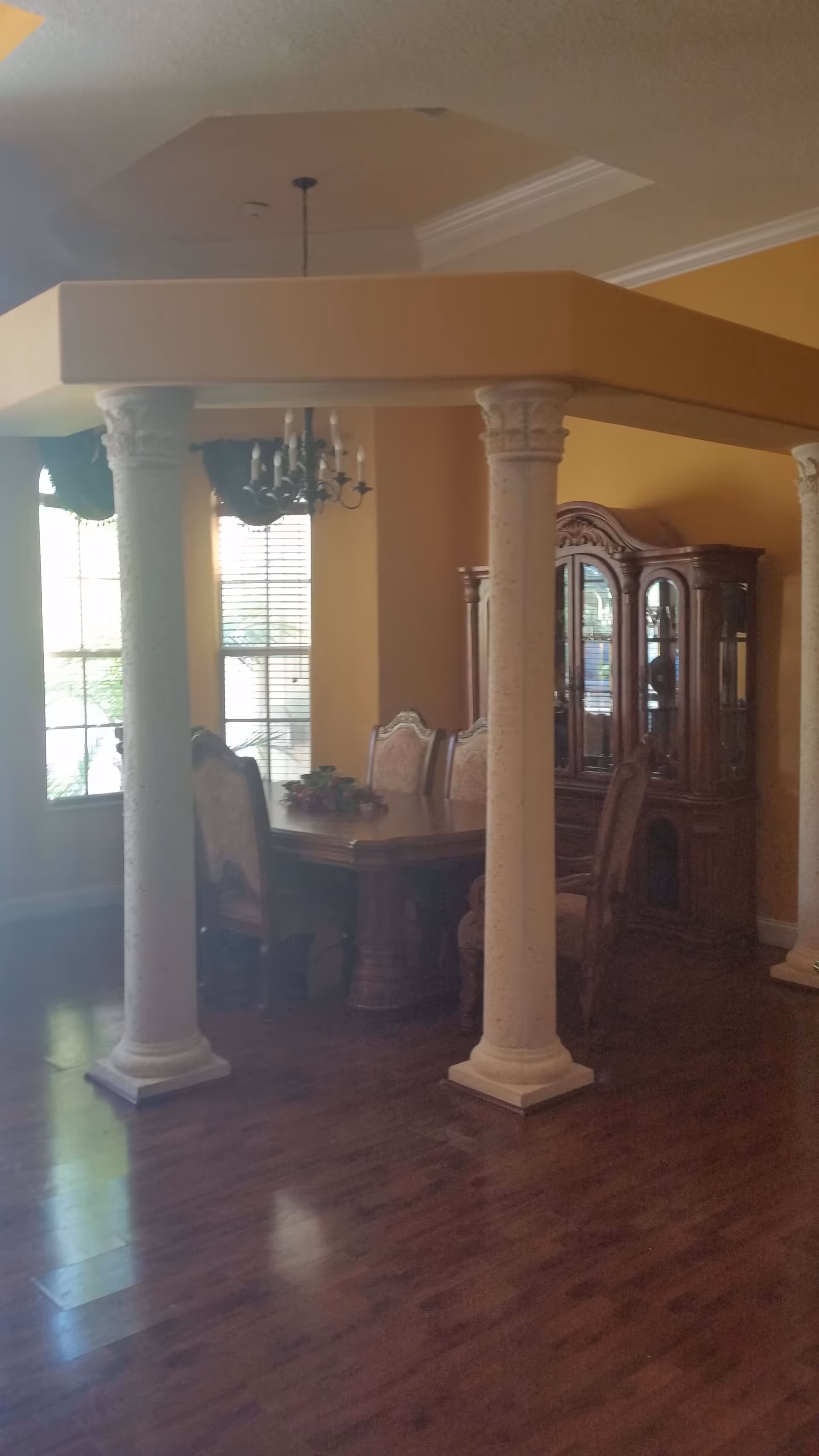 Dining room with decorative columns surrounding a wooden dining table, upholstered chairs, chandelier, and a china cabinet against yellow walls.