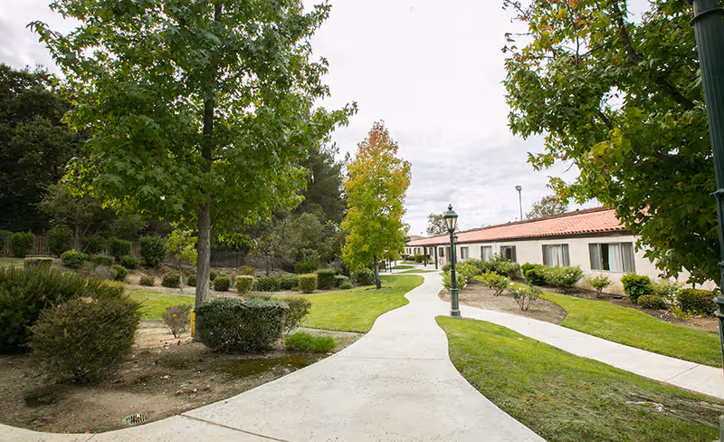 A winding concrete pathway through a landscaped garden area with green grass, bushes, and trees. On the right side, there is a single-story building with a red-tiled roof and multiple windows. Two green lamp posts are visible along the pathway under a cloudy sky.