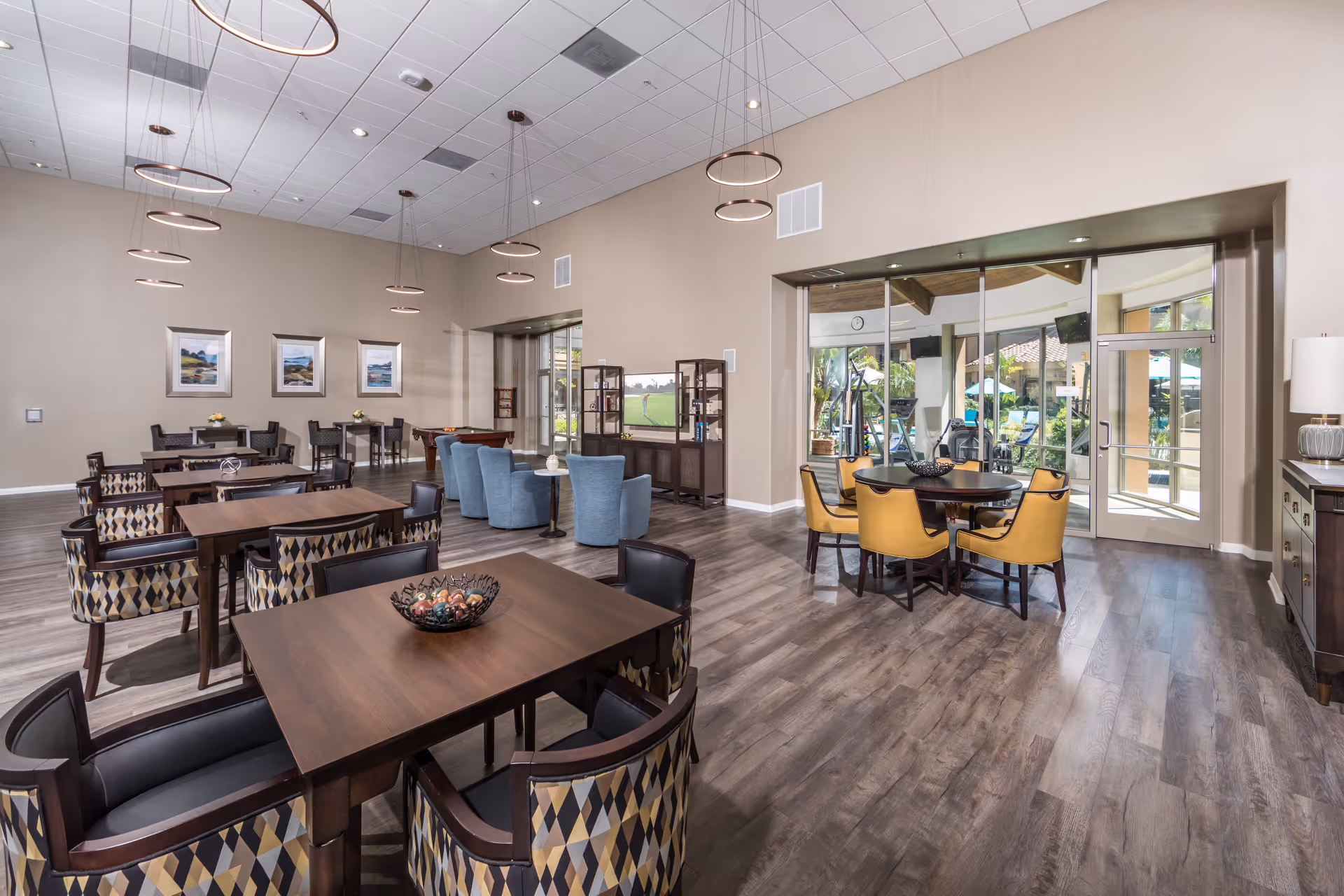 Spacious and well-lit common area with multiple seating arrangements including tables with patterned chairs, a round table with yellow chairs, and blue armchairs facing a television. The room features modern circular pendant lights, wood flooring, and large glass doors leading to an outdoor area with exercise equipment and greenery.