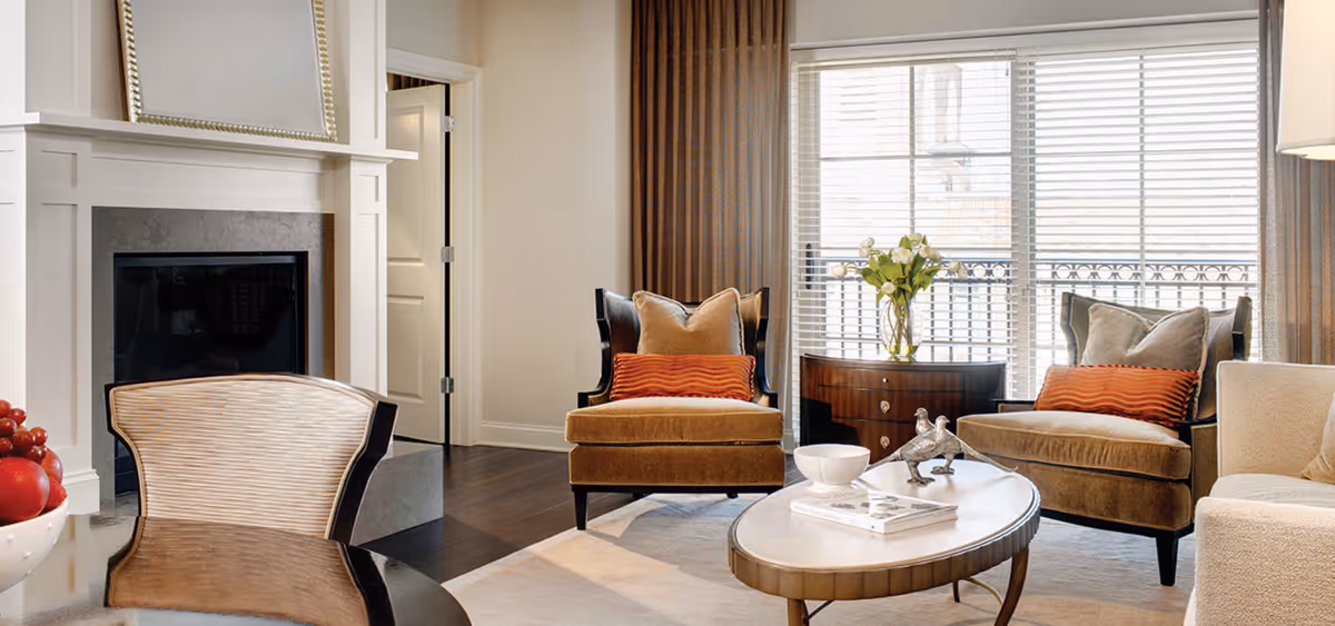 Bright living room with a fireplace, two upholstered chairs with orange pillows, a coffee table and a large window with blinds.