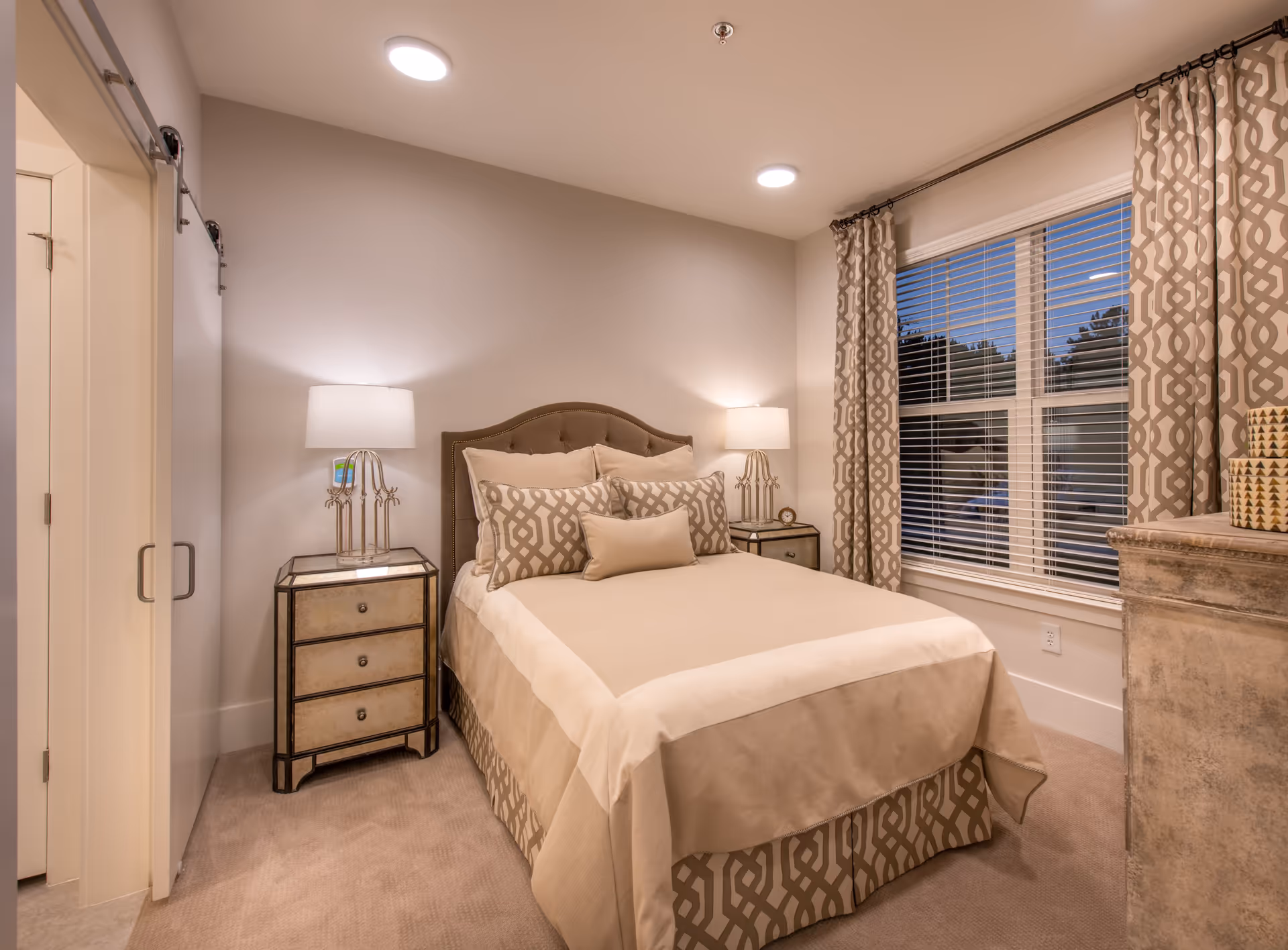 A cozy bedroom with a neatly made bed featuring beige and patterned pillows and bed skirt. Two bedside tables with matching lamps flank the bed. A window with patterned curtains is on the right wall, and a dresser with decorative items is partially visible. The room has soft lighting and a neutral color palette.