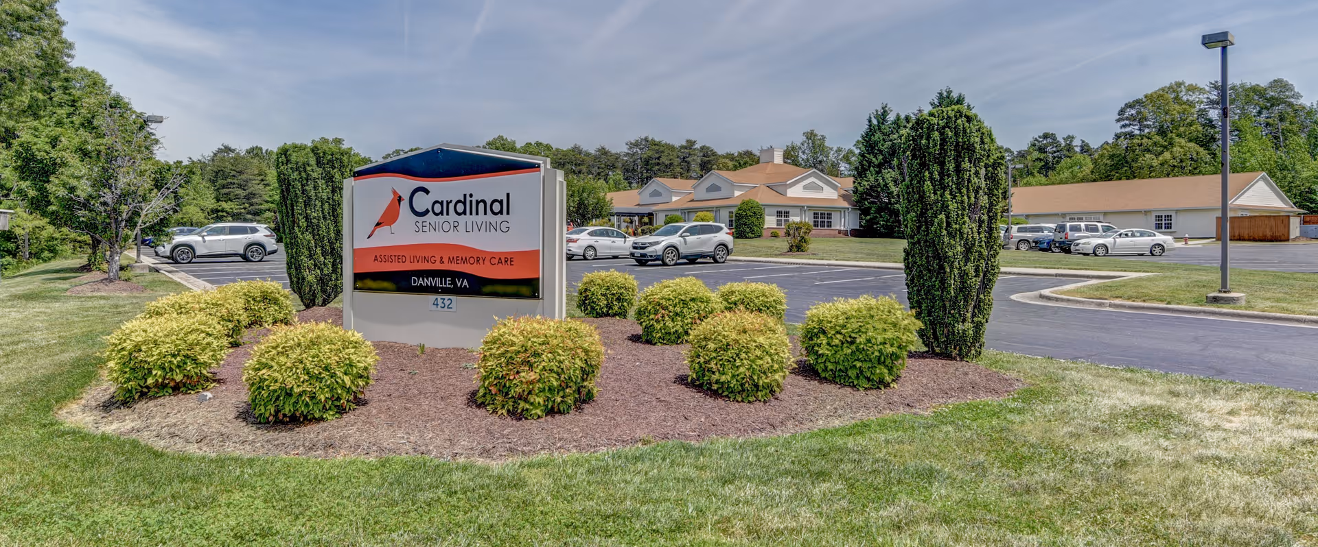 Outdoor view of Cardinal Senior Living at Danville facility sign surrounded by landscaped bushes and grass, with a parking lot and buildings in the background under a clear sky.