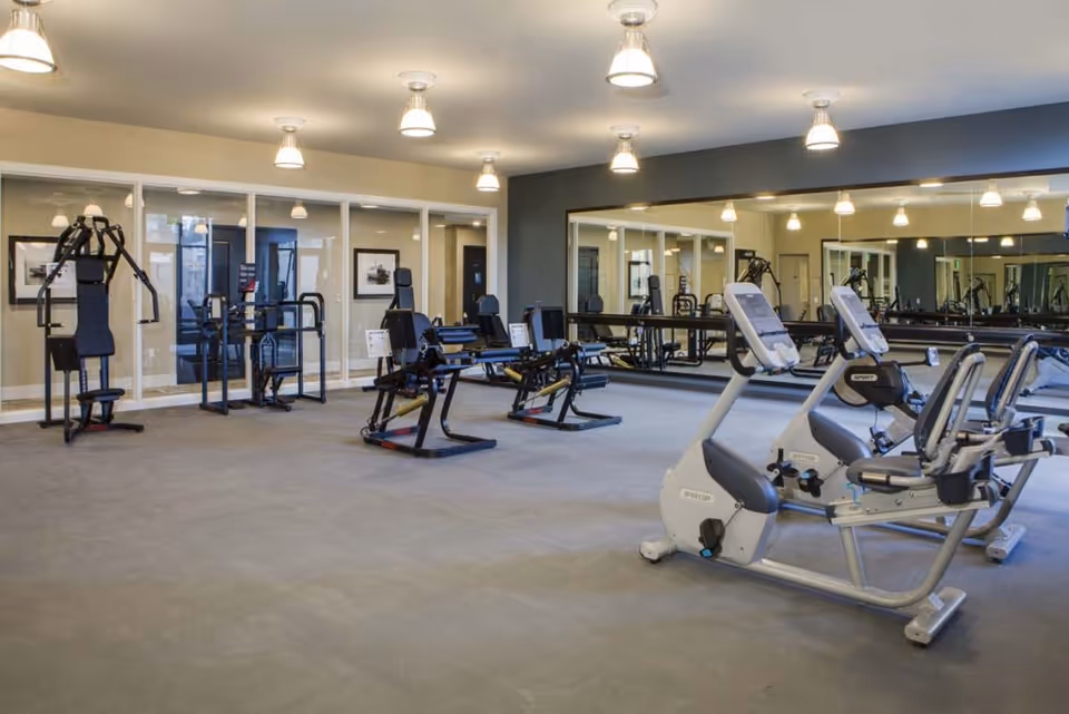 A spacious fitness room with various exercise equipment including stationary bikes, weight machines, and strength training equipment. The room has large mirrors on one wall and glass windows on another, with ceiling lights providing bright illumination.