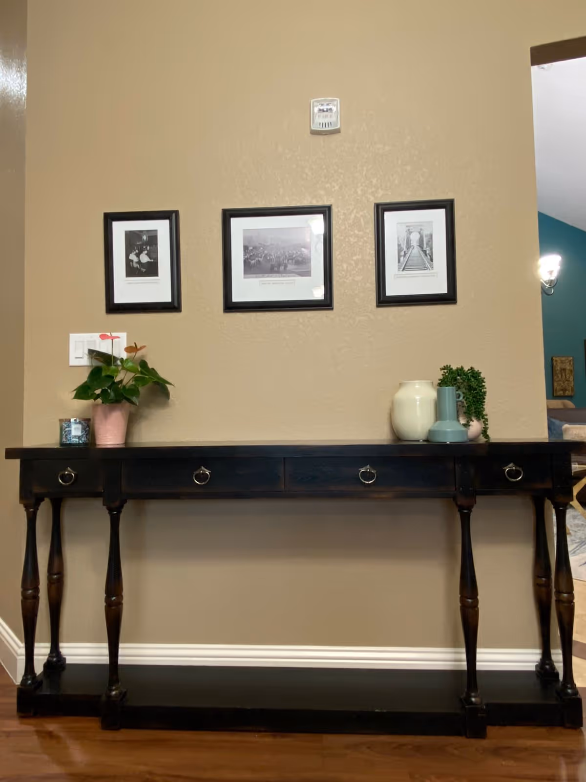 A dark wooden console table with four drawers and turned legs against a beige wall. On the table are decorative items including a potted plant, a candle, a white vase, a blue vase, and a small green plant. Above the table are three framed black and white photographs hung on the wall. A light switch is visible to the left of the table, and an open doorway to another room is on the right.