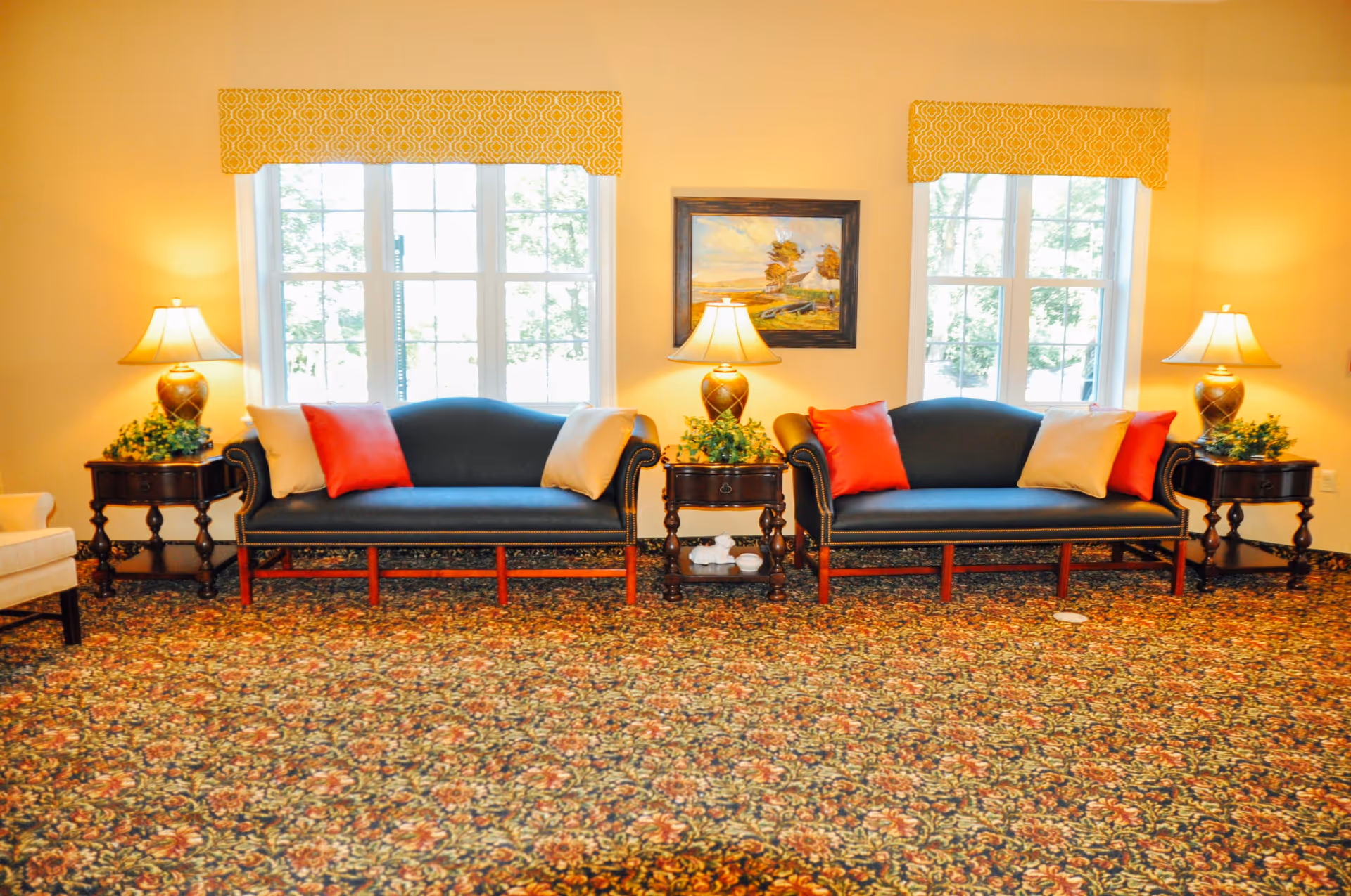 A cozy living room area with two dark blue upholstered sofas, each adorned with red and beige throw pillows. Between the sofas are two wooden side tables with decorative lamps and green plants. The room has large windows with yellow patterned valances, a floral carpet, and a framed landscape painting on the wall.