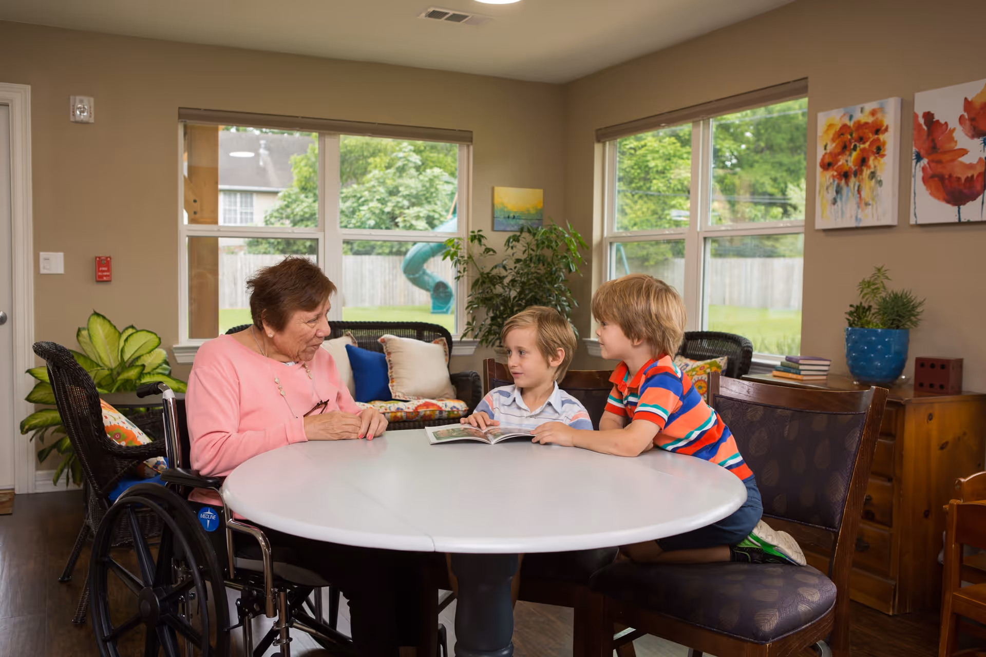 An elderly woman in a wheelchair sitting at a round table with two young boys who are looking at a book together in a bright room with large windows, plants, and colorful wall art.