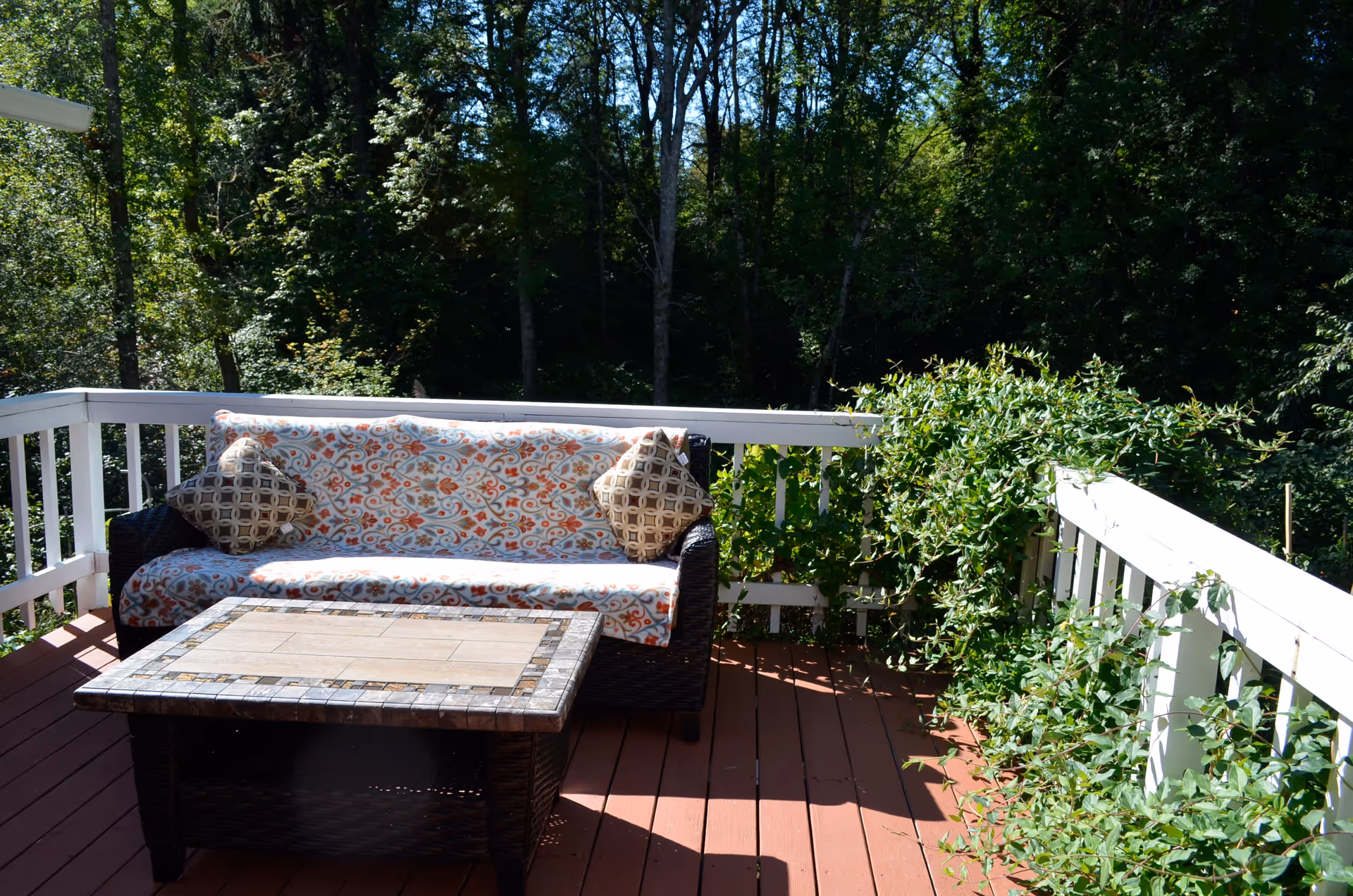 A sunlit wooden deck with a patterned cushioned outdoor sofa and a tiled coffee table beside white railing and leafy trees.