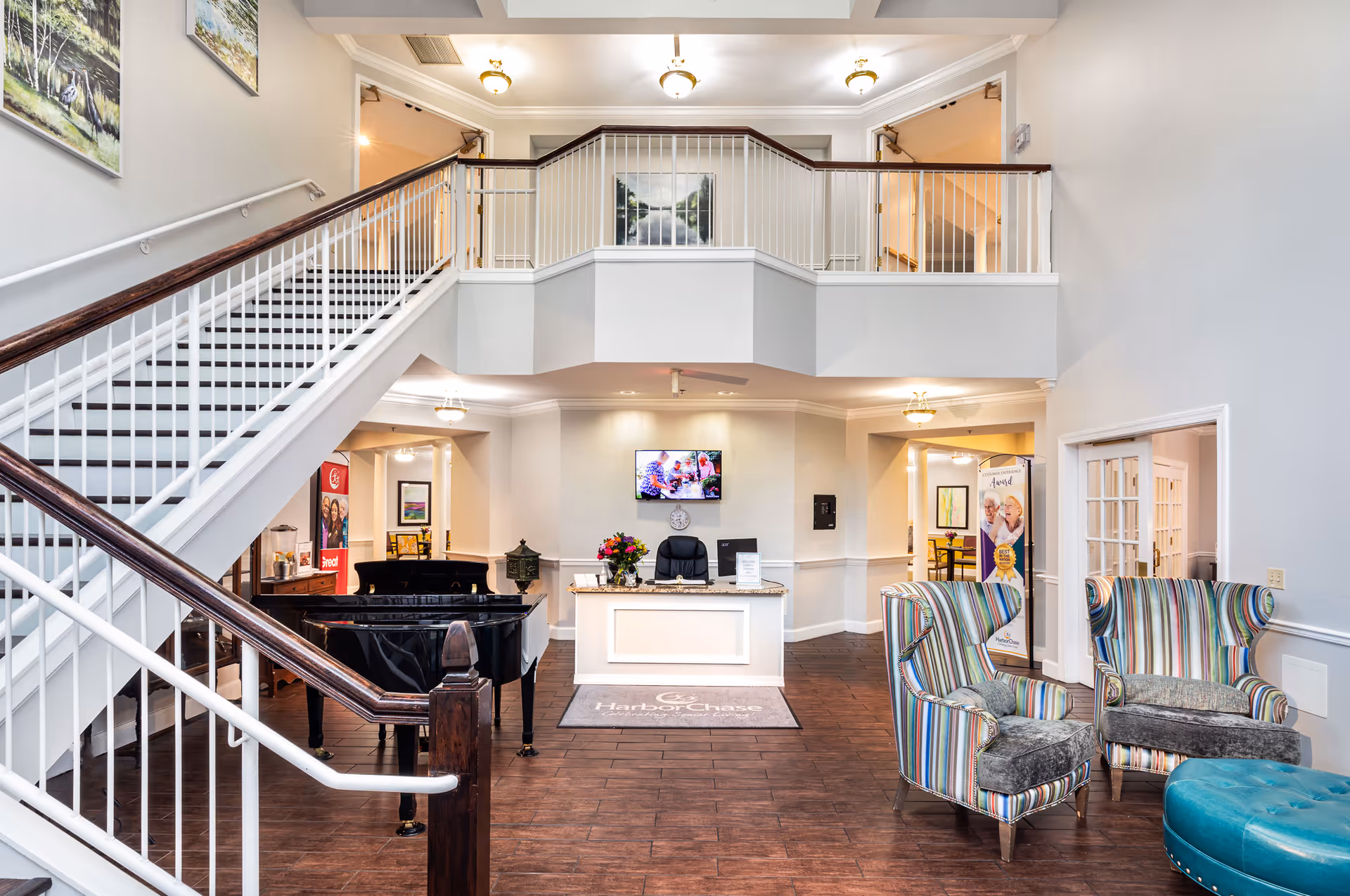 Interior view of a senior living facility lobby with a staircase on the left, a black grand piano, a reception desk with a chair and flowers, two colorful striped armchairs, and a teal ottoman. The walls are light gray with paintings and the floor is dark wood. There is a balcony railing above the reception area and ceiling lights.