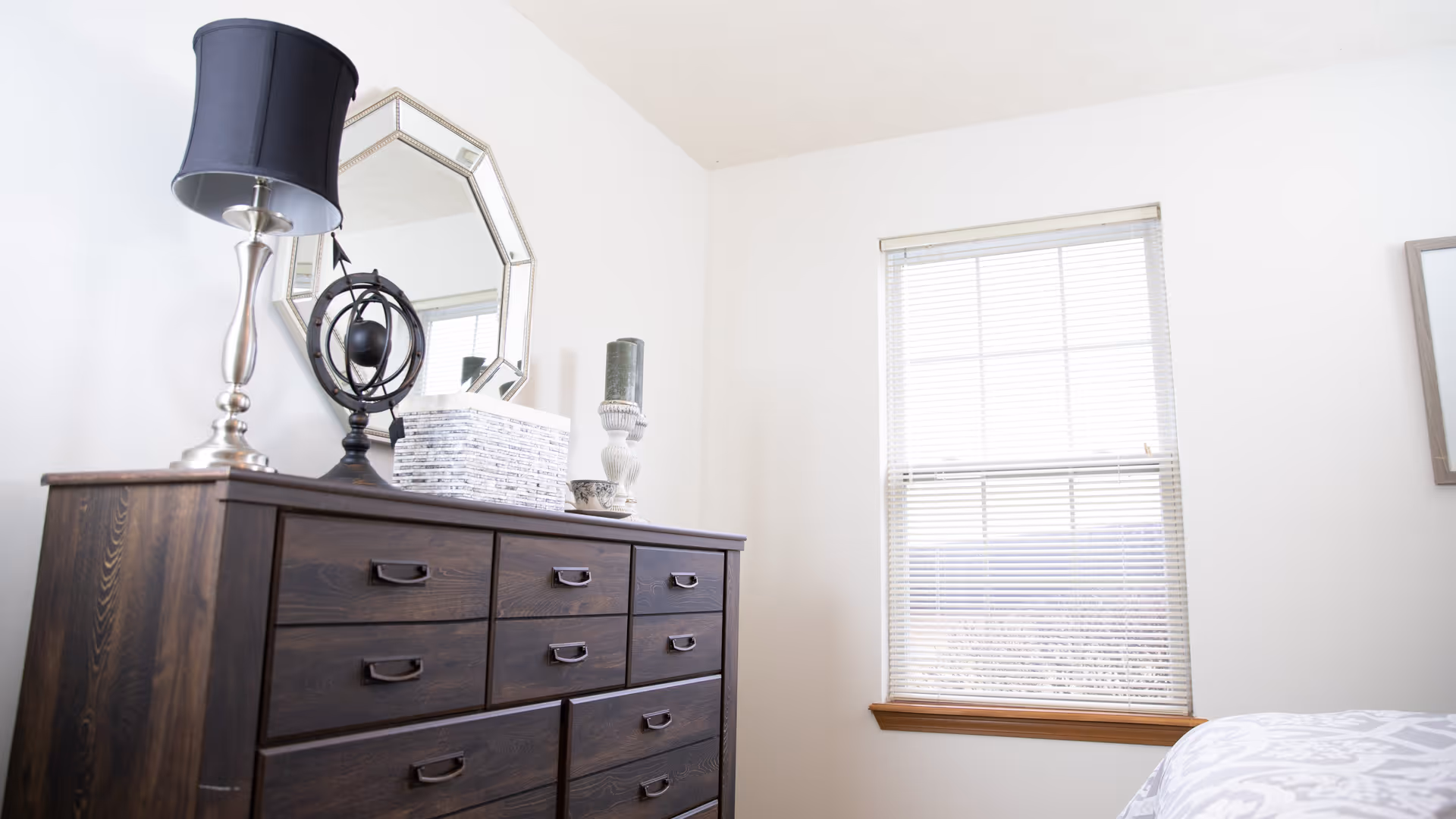 A bright bedroom corner featuring a dark wooden dresser with multiple drawers. On top of the dresser are decorative items including a silver lamp with a black lampshade, a black spherical sculpture, a stack of white books, and a candle holder. A geometric mirror hangs on the white wall above the dresser. To the right, there is a window with white blinds partially open, and part of a bed with a white and gray patterned comforter is visible.