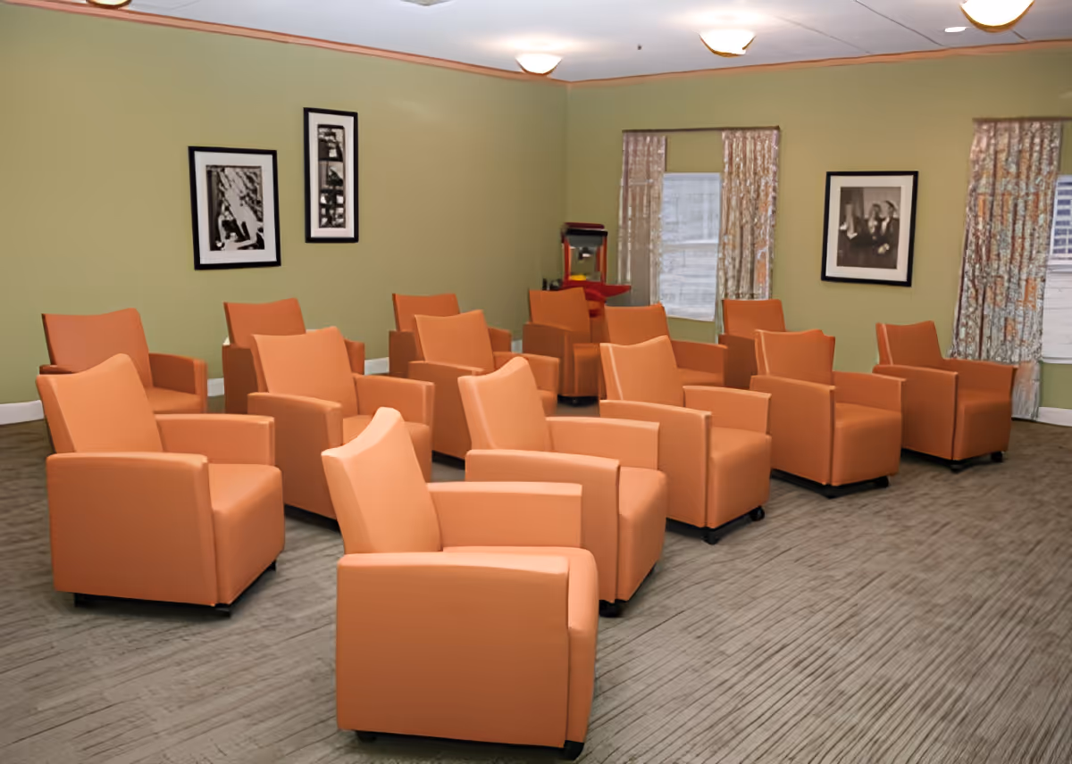 A communal room with multiple orange armchairs arranged in rows, green walls with framed artwork and windows with patterned curtains.