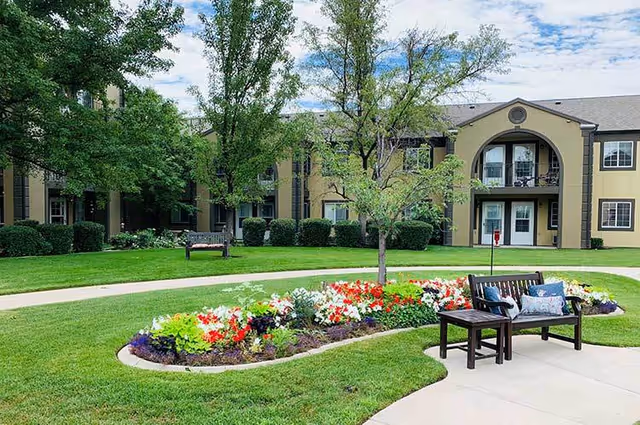 A landscaped outdoor area at Cedarwood at Sandy featuring a curved flower bed with colorful flowers, green grass, a small tree, and a wooden bench with cushions. In the background, there is a two-story building with balconies and windows under a partly cloudy sky.