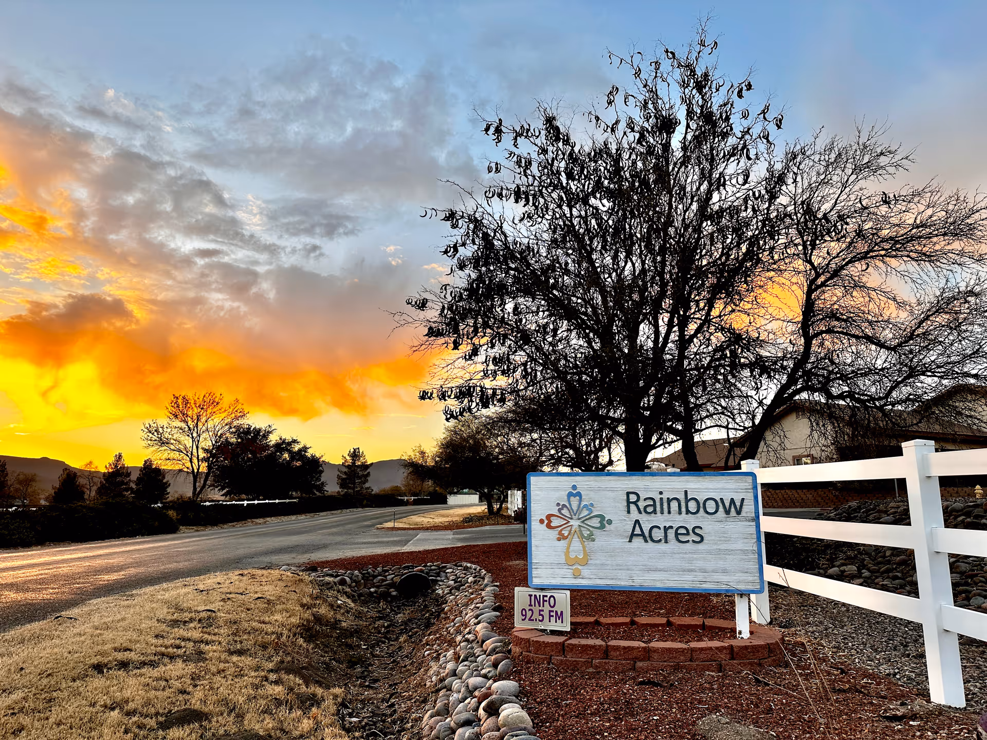 A roadside view at sunset with a sign that reads 'Rainbow Acres' next to a white fence and leafless trees. The sky is partly cloudy with vibrant orange and yellow hues from the setting sun.