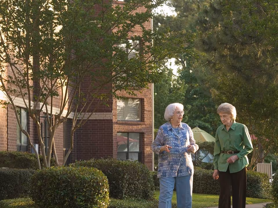 Two elderly women walking and talking outside near a brick building surrounded by trees and bushes in a garden-like setting.