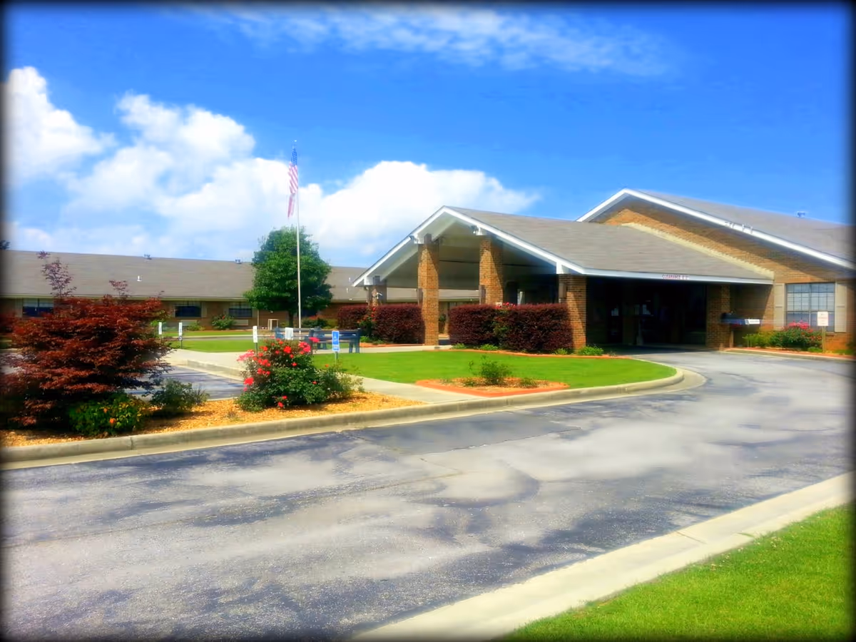 Front exterior of a single-story brick health and rehabilitation center with a covered entrance, driveway, landscaped lawn, and an American flag.