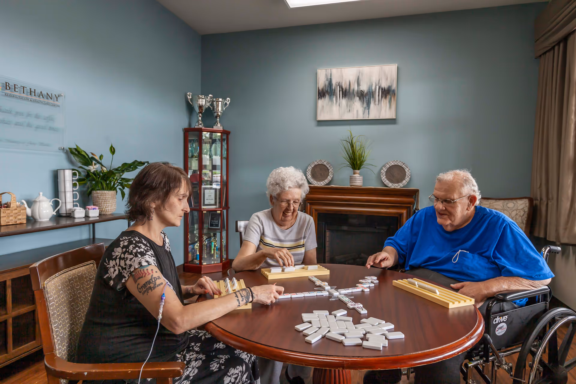 Three people sitting around a round wooden table playing a game of dominoes in a cozy room with blue walls. One person is in a wheelchair, and another has visible tattoos on their arm. Behind them is a cabinet with trophies, a fireplace with decorative plates and a plant, and a side table with a teapot and cups.