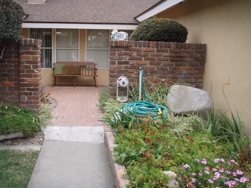 Front entrance of a care home with a brick pathway leading to a wooden bench with green cushions. The entrance is flanked by low brick walls and landscaped garden beds with green plants, flowers, and a coiled green garden hose. The house number 1927 is displayed above the brick wall.