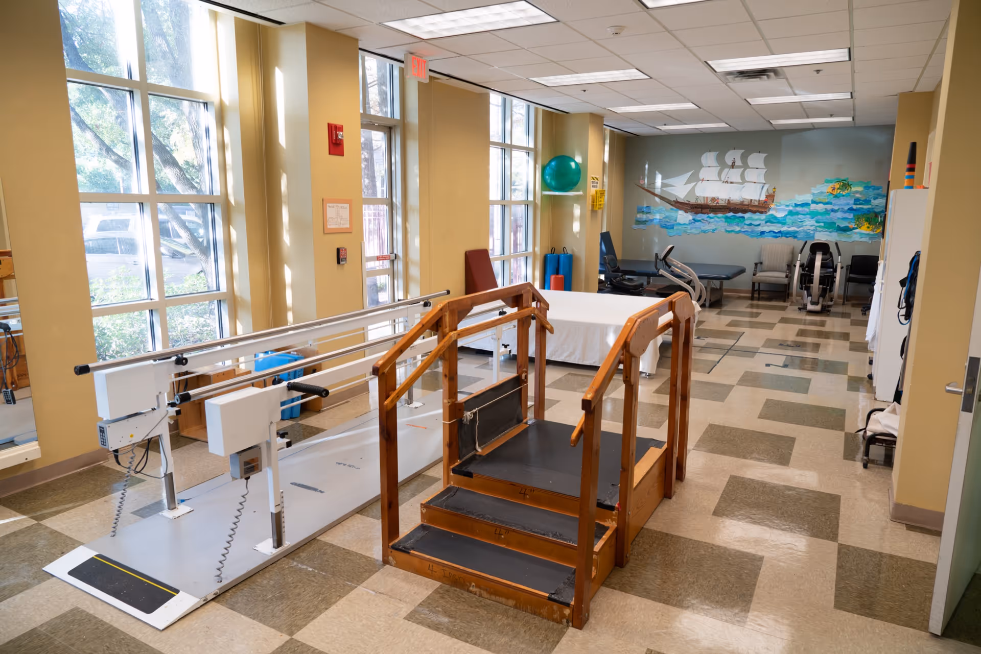 A physical therapy room with parallel bars for walking exercises, a small wooden staircase with handrails, exercise equipment, chairs, and a mural of a sailing ship on the wall. Large windows let in natural light.