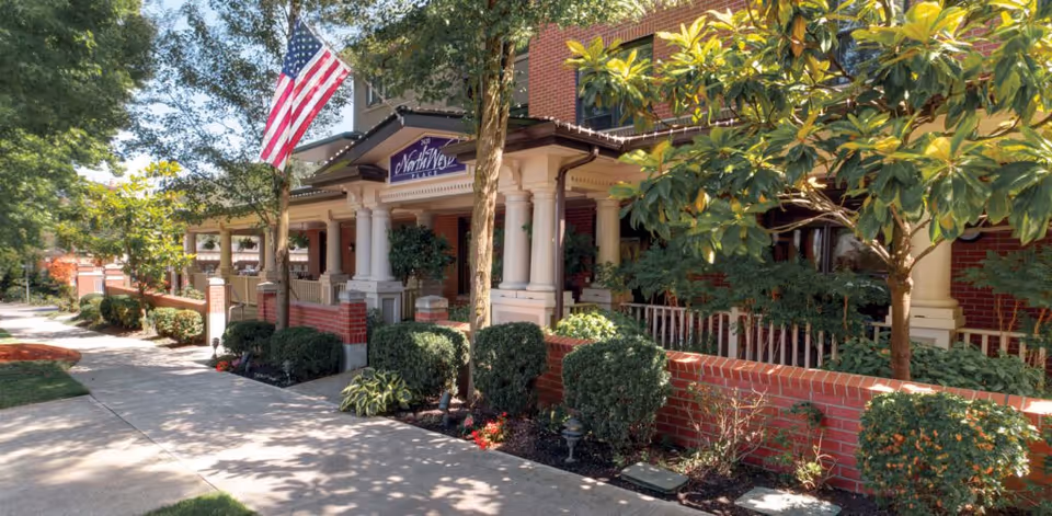 Exterior view of NorthWest Place senior living facility showing a brick building with a covered porch supported by white columns, an American flag, and well-maintained landscaping including bushes and trees along a sidewalk.