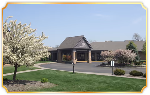 Exterior view of The Inn At Belden Village building with a driveway, landscaped lawn, and blooming trees under a clear blue sky.