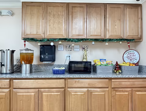 A kitchenette with wooden cabinets, a coffee urn, microwave, and snack baskets along a counter.