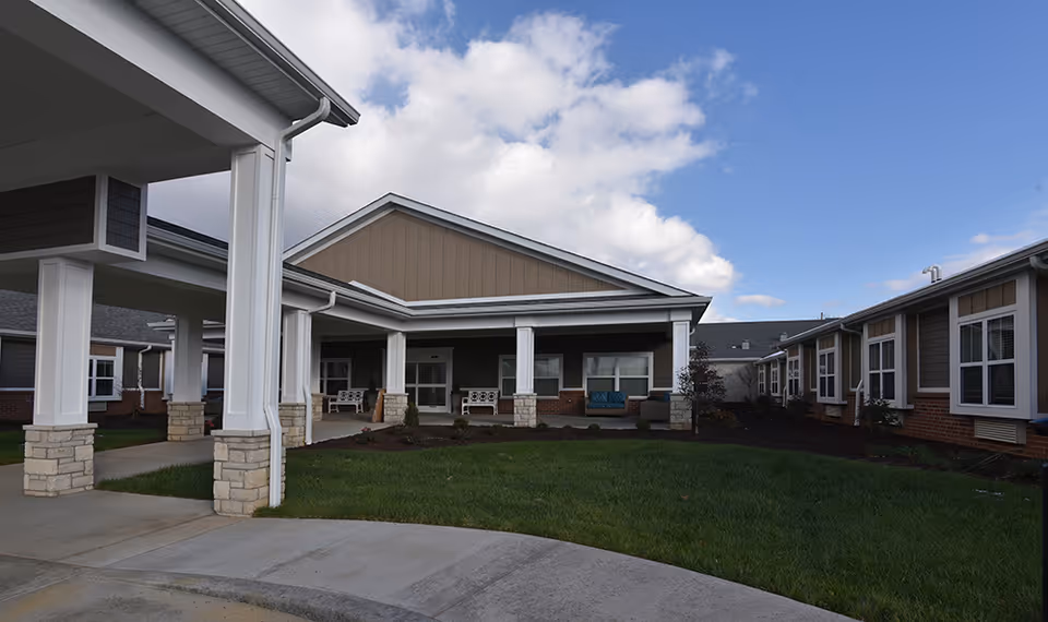 Exterior view of a senior living facility with a covered entrance supported by white columns with stone bases, a green lawn in front, and a partly cloudy sky above.