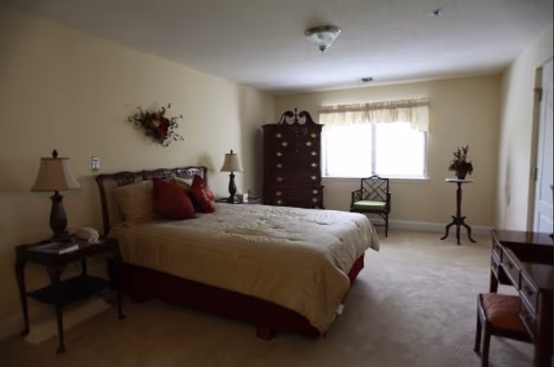 A bedroom with a large bed covered in beige bedding and red pillows. There are two bedside tables with lamps on either side of the bed. A tall wooden dresser is positioned against the far wall near a window with sheer curtains. A wooden chair with a green cushion and a small round table with a floral arrangement are also visible. The walls are painted light beige and the floor is carpeted.