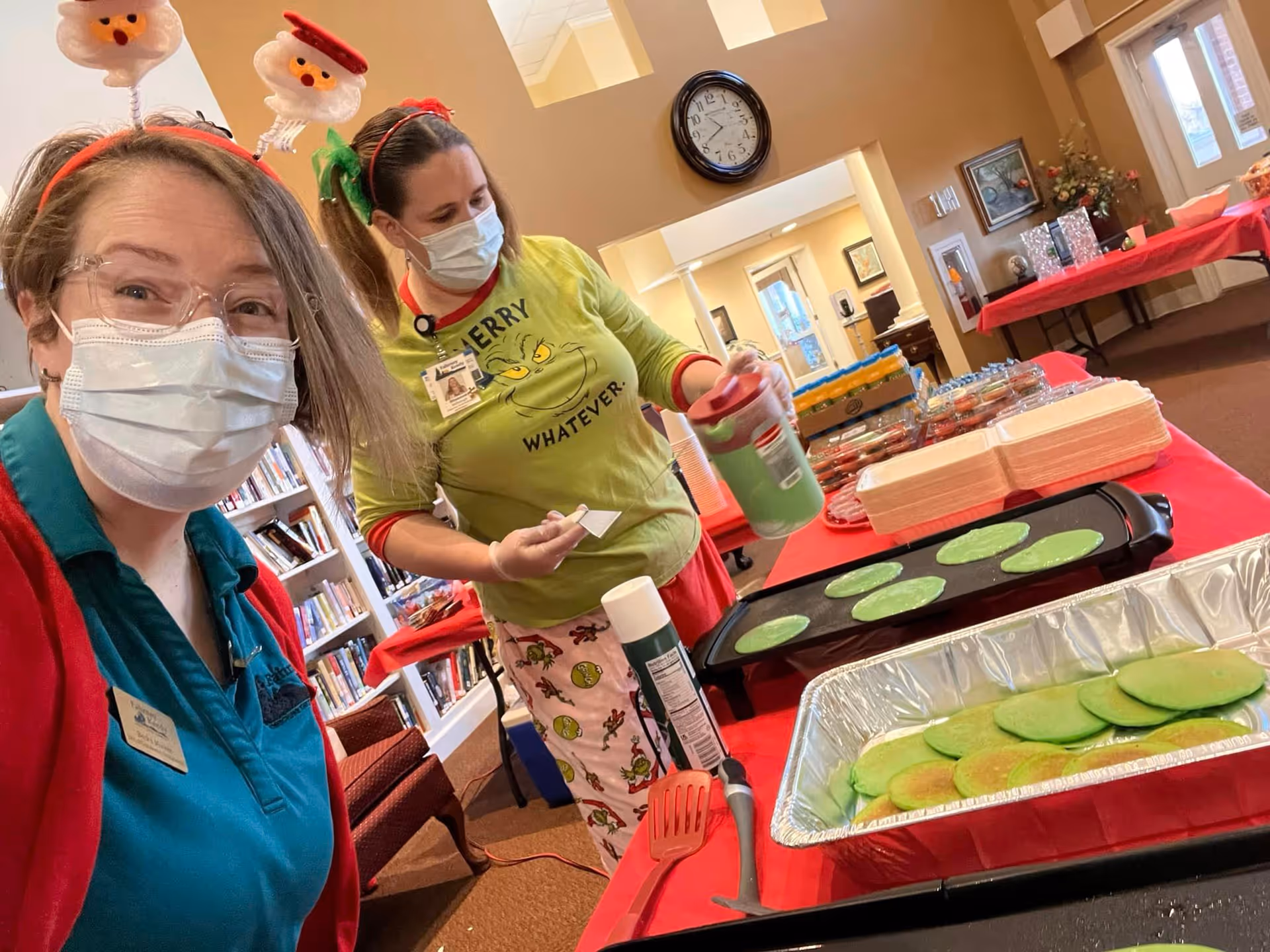 Two women wearing face masks and festive headbands are preparing green pancakes on a griddle in a room with bookshelves and tables covered with red tablecloths. One woman is smiling at the camera while the other is pouring pancake batter. The setting appears to be a community or senior living facility.