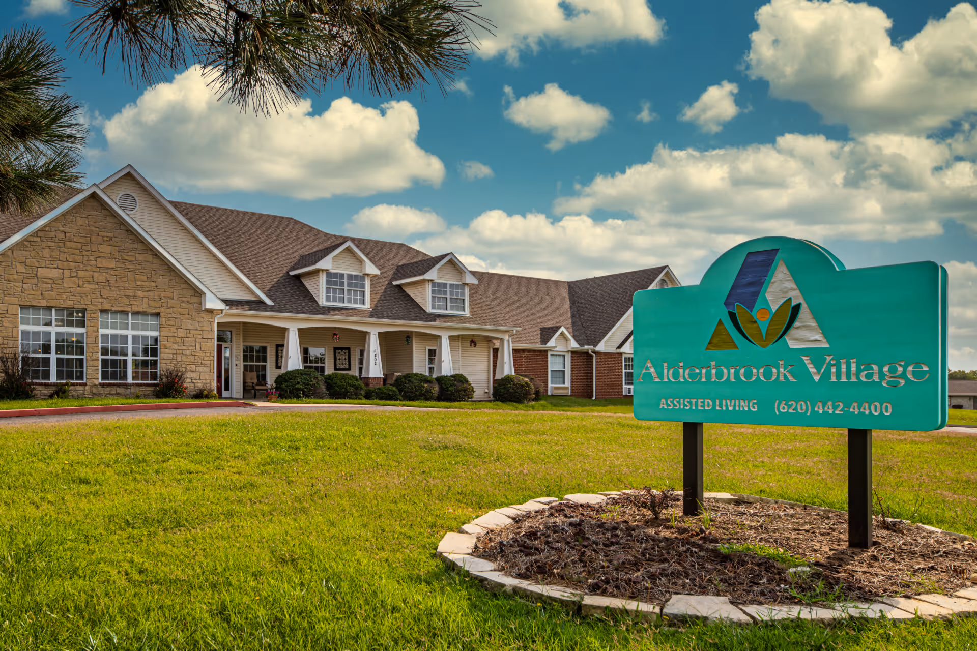Exterior view of Alderbrook Village assisted living facility with a large green sign displaying the facility name and phone number in the foreground, a well-maintained lawn, and a building with stone and siding exterior under a partly cloudy sky.