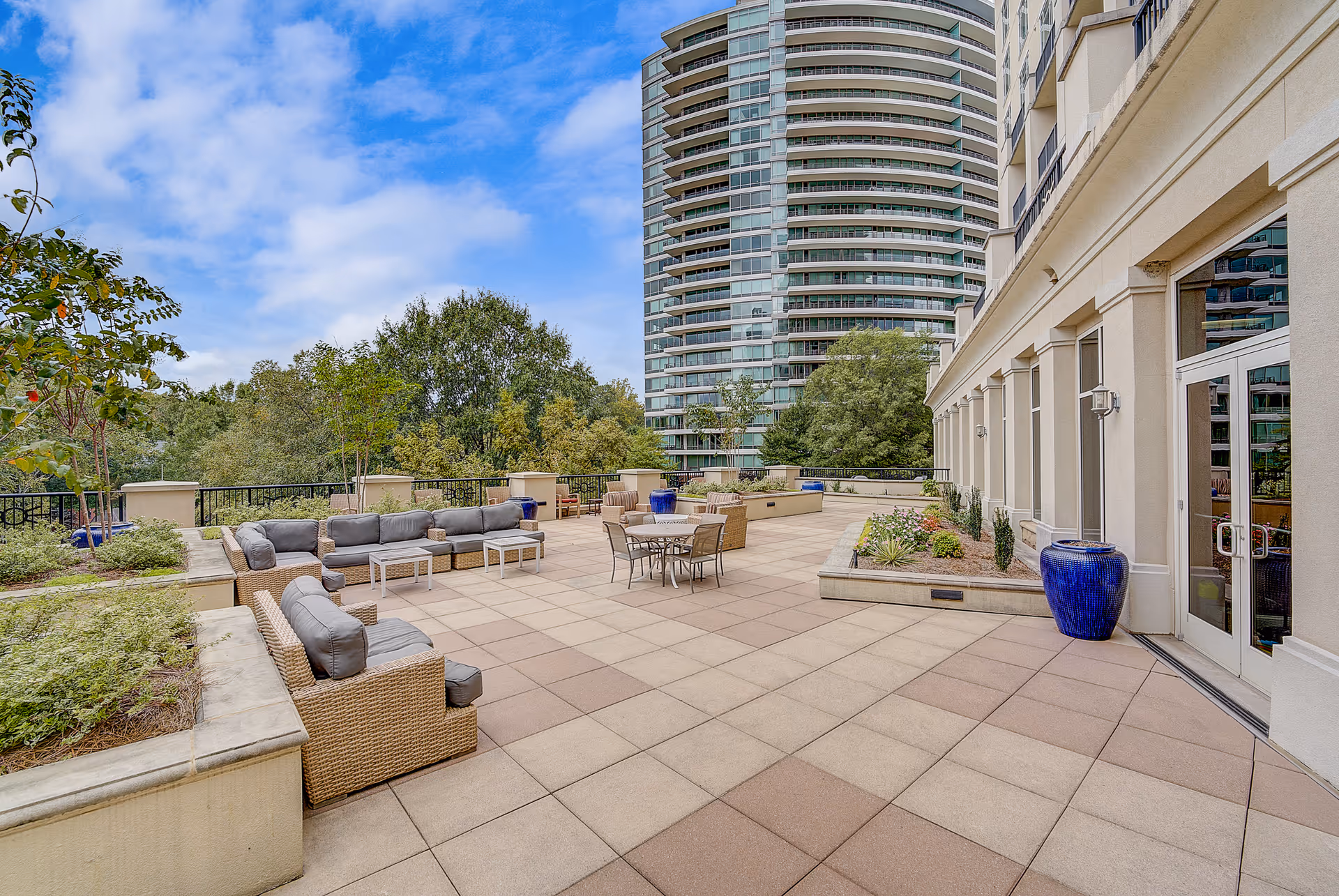 Spacious outdoor patio area with cushioned wicker seating, tables, and chairs. The patio is surrounded by planters with greenery and large blue decorative pots. In the background, there are tall modern buildings and trees under a partly cloudy sky.