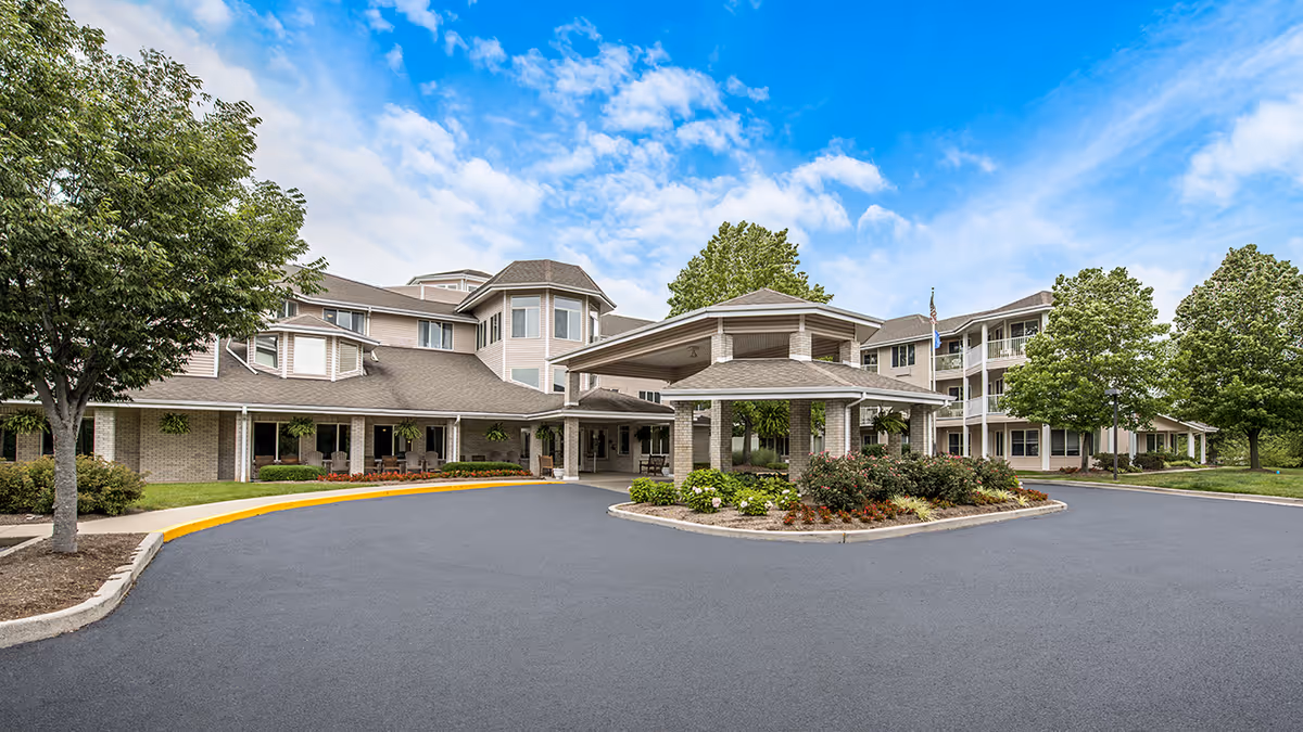 Front exterior of Holiday Redbud Hills senior living building with a porte-cochère, circular driveway, landscaped island, and trees beneath a blue sky.