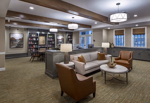 A spacious and well-lit senior living common area featuring a beige sofa with brown cushions, two brown leather armchairs, a round coffee table, and two table lamps. In the background, there are bookshelves filled with books and decorative items, a table with chairs, and large windows with brown blinds. The ceiling has wooden beams and modern light fixtures.