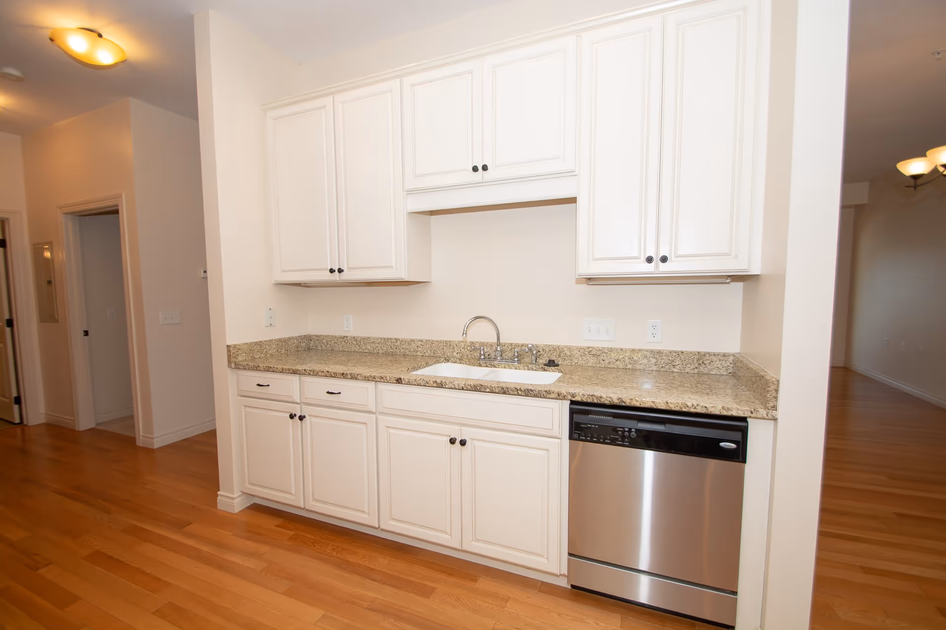 A kitchen area with white cabinets, granite countertops, a stainless steel dishwasher, and a white sink with a chrome faucet. The floor is wooden, and there are doorways visible leading to other rooms.