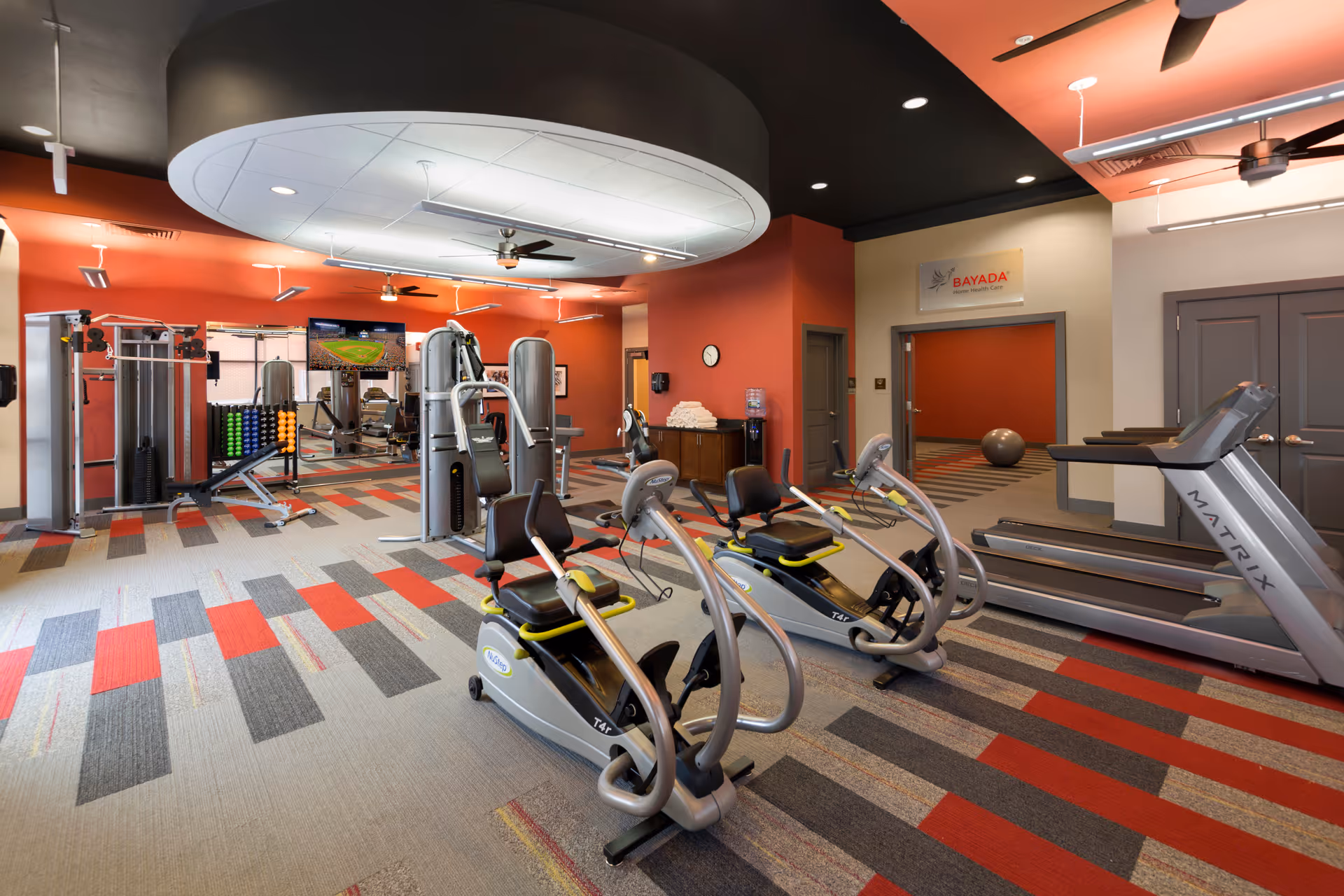 A brightly colored fitness room featuring cardio machines, strength equipment, and patterned carpet flooring.
