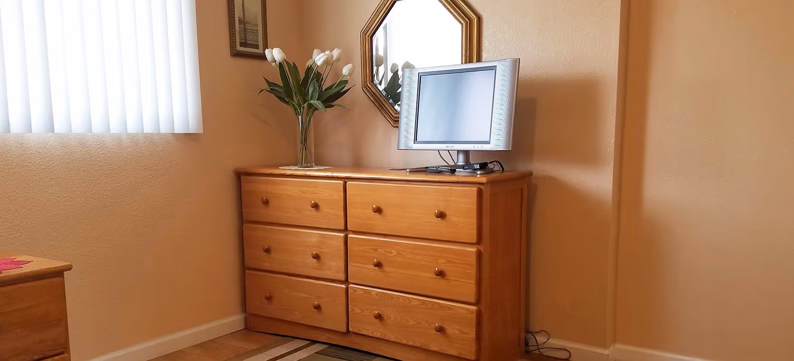 A corner of a bedroom featuring a wooden dresser with six drawers, a small flat-screen TV on top, a vase with white tulips, and a decorative mirror hanging on the wall above. The room has beige walls, a window with vertical blinds, and a portion of a wooden bed frame visible on the left.