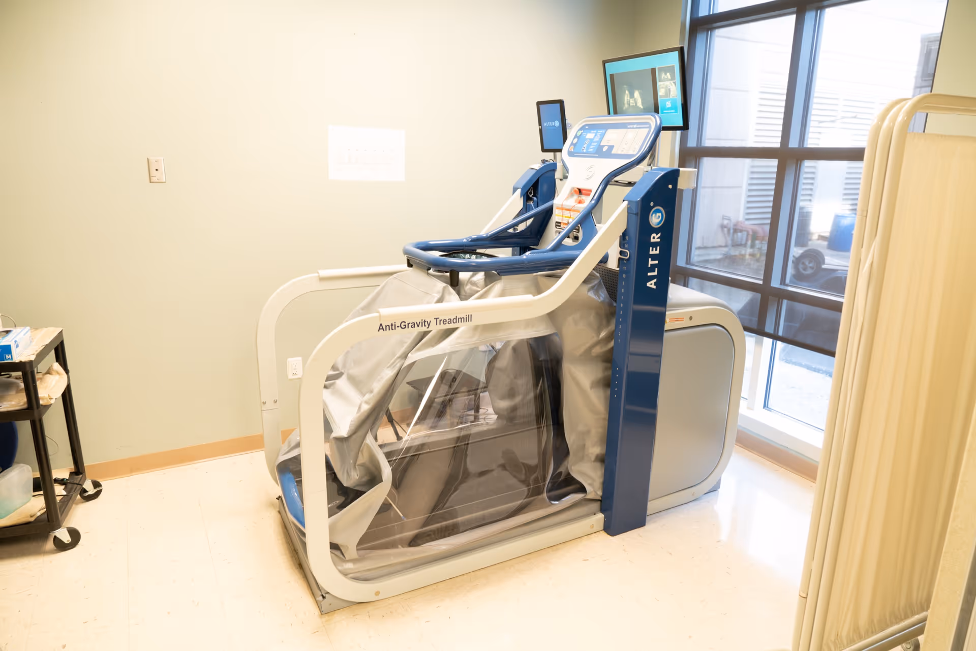An anti-gravity treadmill machine in a room with light-colored walls and a large window. The treadmill has a clear plastic enclosure and digital screens. There is a small cart with supplies on the left side and a privacy curtain on the right.