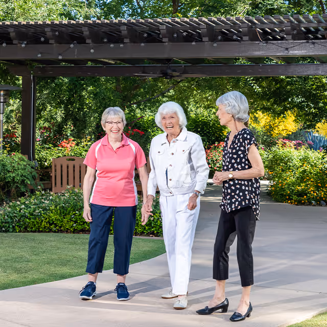Three elderly women standing and smiling outdoors in a garden area with greenery and flowers around them. They are under a wooden pergola with string lights, enjoying a sunny day at Rosewood Retirement Community.